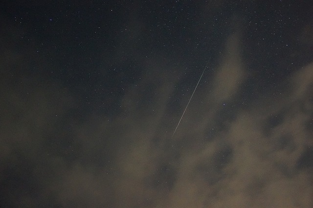 薄雲の広がる星空の中央やや右に、右上から左下にかけて画面の3分の1ほどの長さの流星が白い筋として見える写真