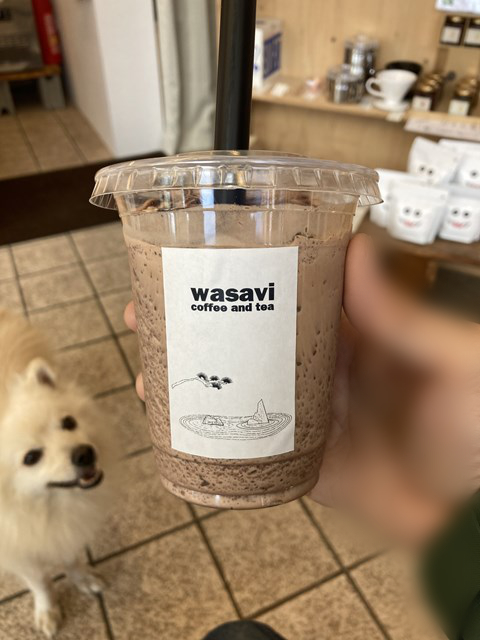 Foto de una bebida marrón en un vaso de plástico con tapa. Un perro blanco está mirando desde el suelo