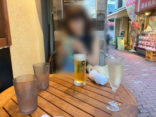 ビールとスパークリングワインが置かれた丸いテーブルの向こうに座る人の写真。その人におねだりしている白いイヌの頭が、テーブルの向こうにちょこんとのぞいています