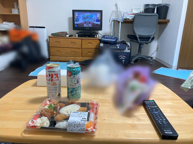 弁当と飲み物が置かれたテーブル越しにテレビが見える写真