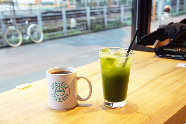 左に茶色の飲み物が入ったマグカップが、右に緑色の飲み物が入ったガラスのコップが写った写真。向こうに線路が見えています